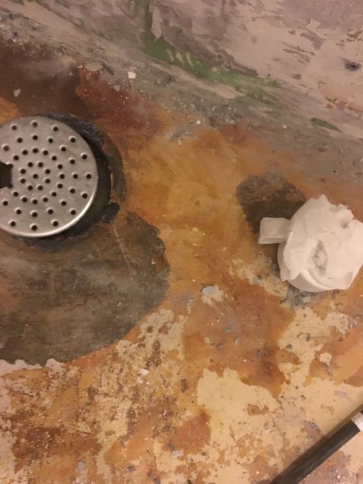 Skadat badrumsgolv med synlig misstänkt fuktskada runt avloppsbrunnen och rester av lim eller färg.