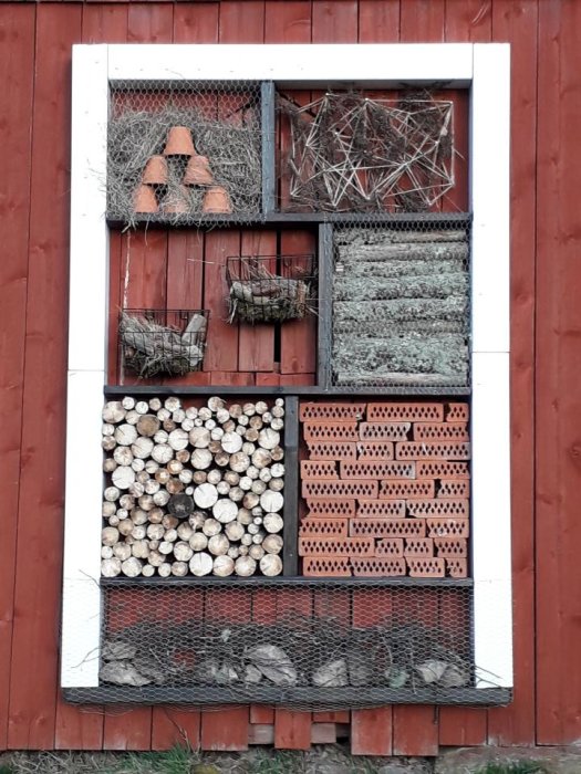 Handgjort insektshotell av spillvirke med olika fack, monterat på en röd trävägg.