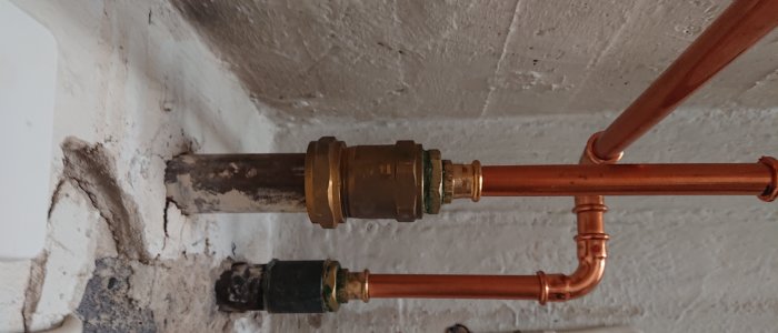 Koppling mellan gammalt rostigt järnrör och nyare kopparledningar med mässingspresskoppling mot vit vägg.