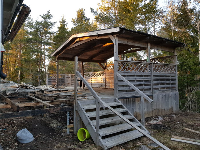 Altan under rivning med kvarstående takkonstruktion och ruttet trägolv och räcken omgivna av skog.