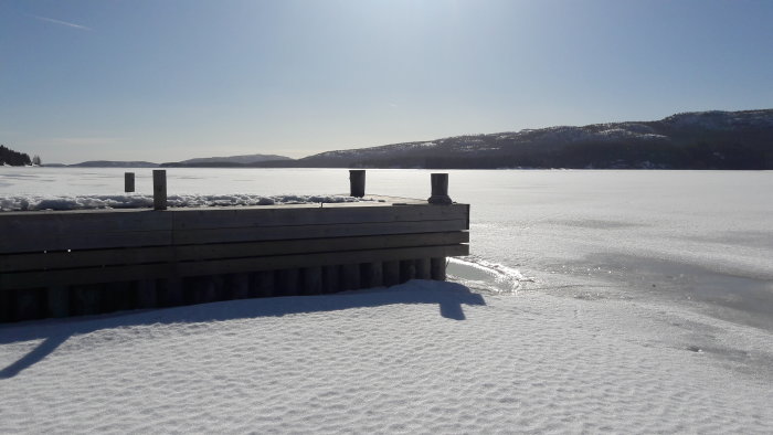 En brygga som sträcker sig över en snötäckt och isbelagd sjö med skogklädda kullar i bakgrunden.