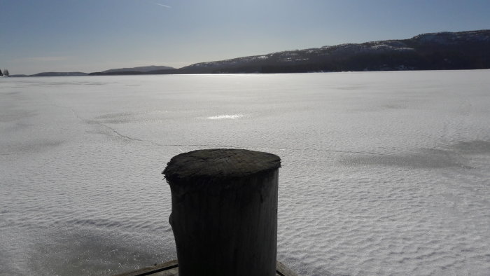 Solbelyst frusen sjö med snö och en bryggas hörn i förgrunden, kulliga landskap i bakgrunden.