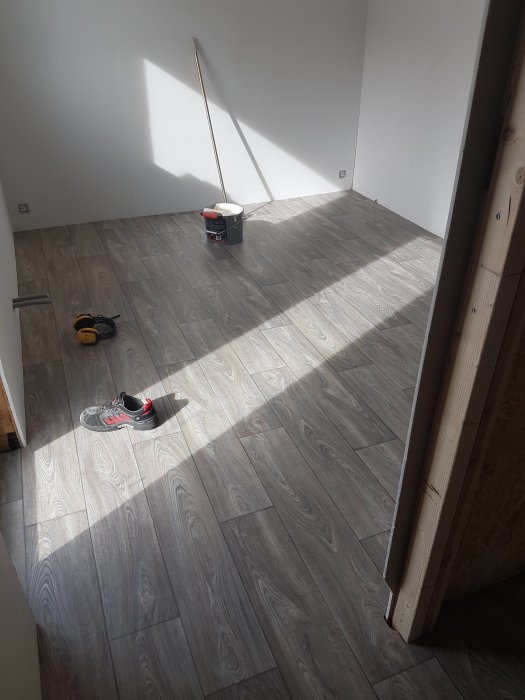 Nybelagd laminatgolv med spackelhink och verktyg, soligt rum under renovering med pardörrar.