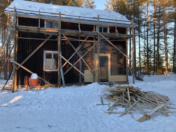 Renoveringsarbete på ett gammalt trähus med ställningar, omgiven av snö och en hög med träsplint.