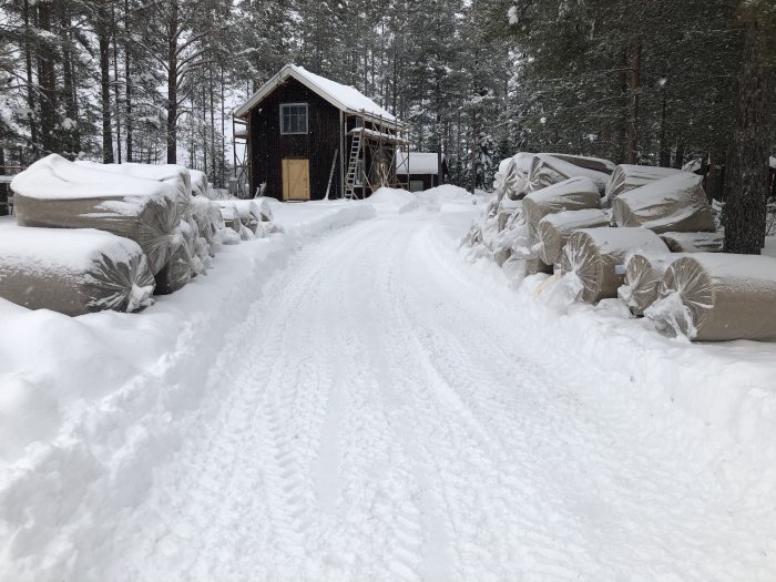 Snötäckt byggarbetsplats med insvepta material och en byggnad i bakgrunden omgiven av skog.