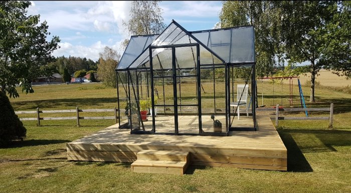 Ett växthus monterat på en träplattform med grön gräsmatta och landskap i bakgrunden.