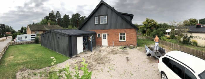 Panoramavy över en pågående renovering av en uppfart utanför ett tegelhus med en person och en släpvagn.