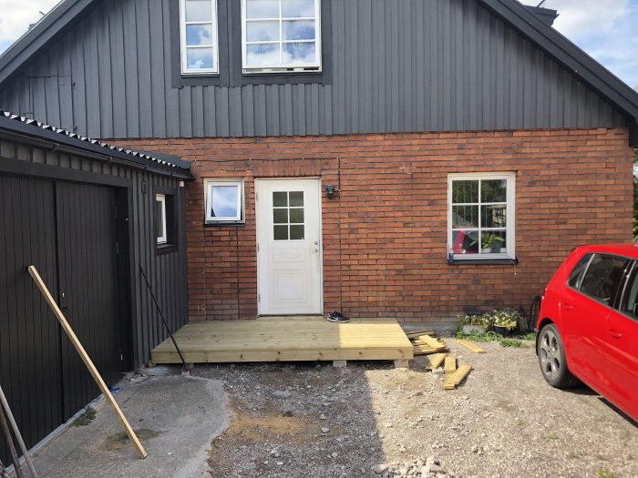 Entré till en villa med oavslutad trätrall utan tak, bredvid garage och en röd bil.