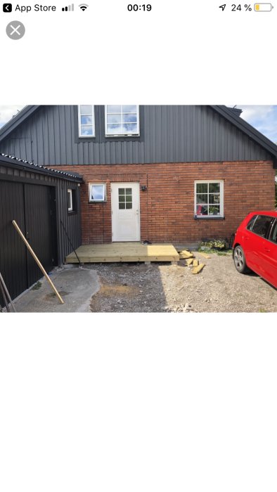 Entré till villa med träveranda, röd tegelvägg, vit dörr och fönster, intill garage med sadeltak.