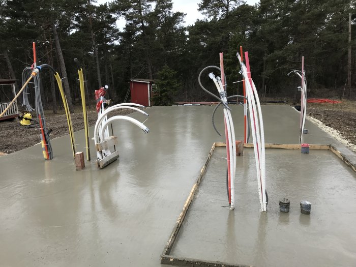 Nygjuten betongplatta med rörledningar och träramar, omgiven av byggmaterial och träd.