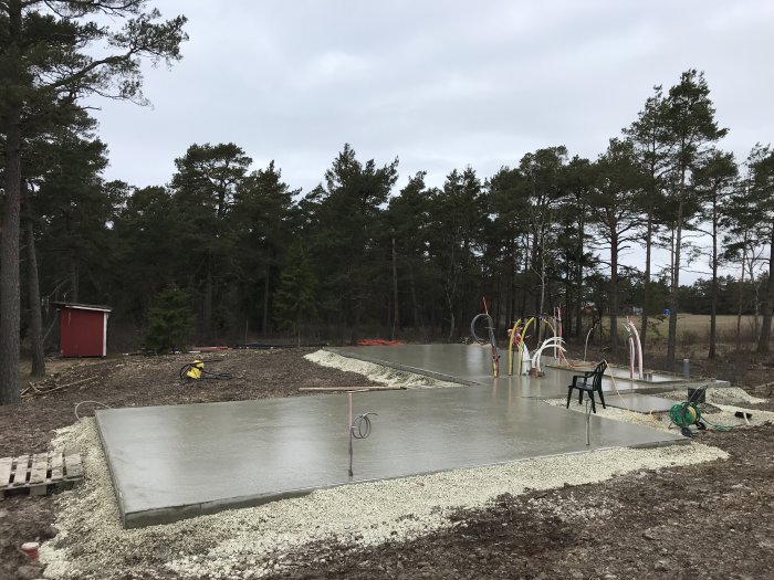 Ny gjuten betongplatta med rör och utrustning i omgivning av natur och röd stuga.