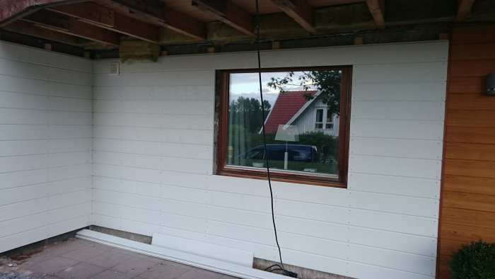 Delvis renoverad husfasad med ny vit panel och ett fönster som speglar ett rött hus och en bil.