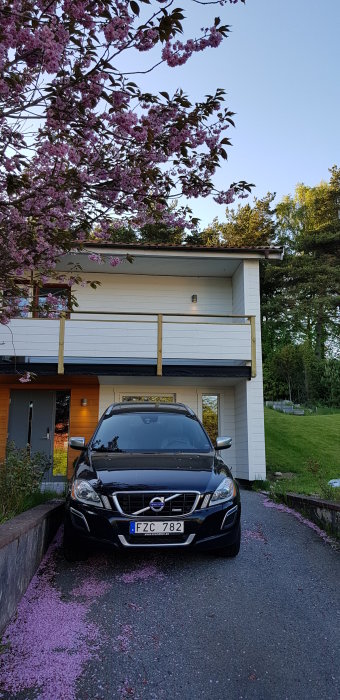 Husets framsida med en pågående balkongkonstruktion, omgiven av blommande träd och en parkerad bil.