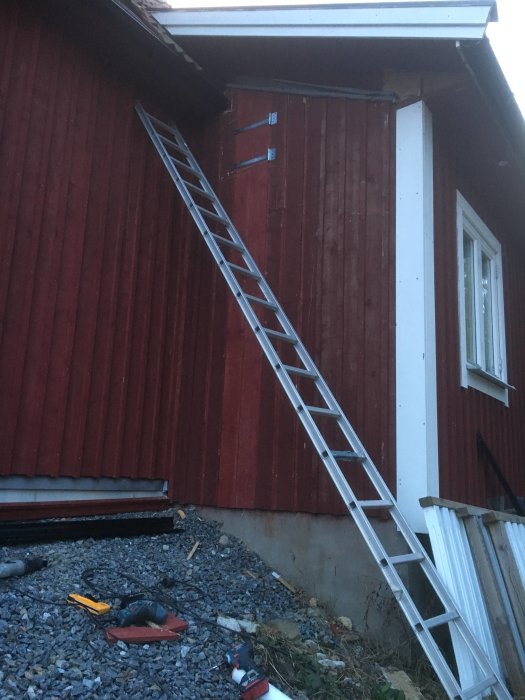 En stege som lutar mot en röd trävägg med öppningar för ventilationsluckor, omgivet av verktyg och byggmaterial.