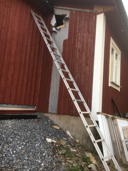 Två öppningar uppkapade i en röd husvägg med en stege lutad mot väggen för installationsarbeten.