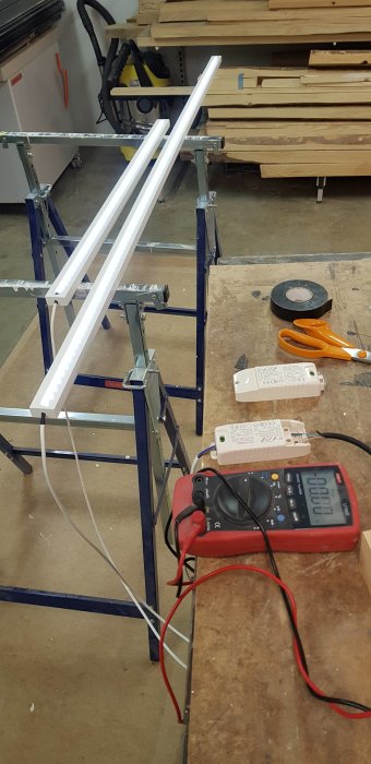 Två LED-listor med tända lampor testas med ett multimeter och konstantströmsdrivdon på ett arbetsbord.