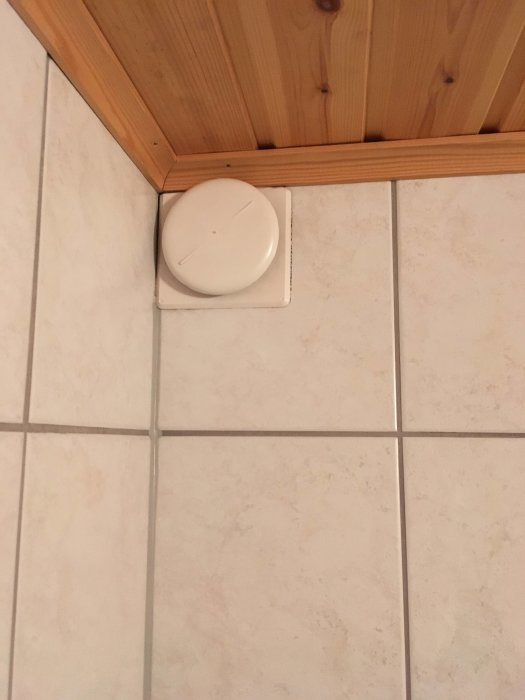 Ventilationsgaller i hörnet av ett kaklat rum med träkant, ser ut som en toalett.
