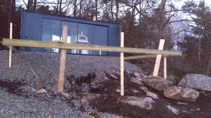 Ombyggd blå container som skall användas som utekök med en påbörjad altankonstruktion och stora stenar placerade framför på en kuperad tomt.