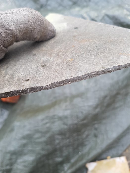 Kädessä pidetään harmaata, kuitumaista levyä, joka voi olla asbestia. Taustalla näkyy peitteinen maa tai rakennelma.