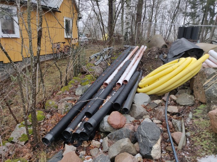 Staplar med oanvända dräneringsrör i olika storlekar och färger förvarade utomhus nära ett gult hus.