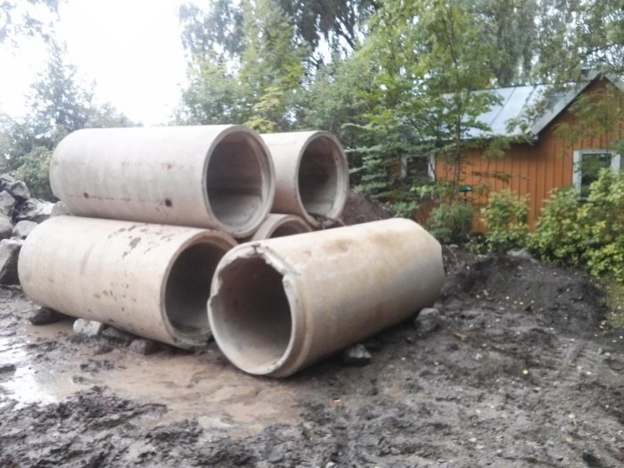 Betonrör ligger på en lerig byggarbetsplats med ett trähus och grönska i bakgrunden.