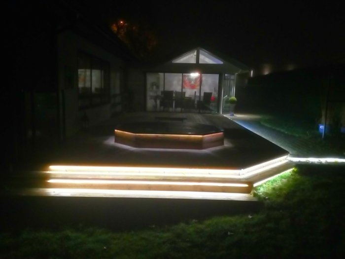Kvällsbild av en trädgård med PLEJD-styrd belysning som lyser längs trappsteg och en terrass, med ett hus i bakgrunden.