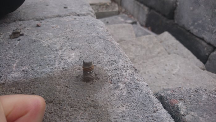 A worn and rusted outdoor expansion bolt in a concrete surface, possibly needing anti-rust treatment.