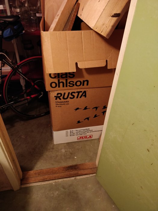 Clas Ohlson och Rusta flyttkartong i hörn med träbitar, bredvid en cykel, i ett garage eller förrådsutrymme.