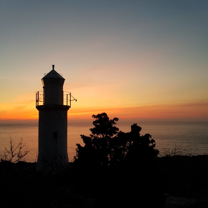 Fyr mot en dramatisk solnedgång över havet med silhuetter av träd.