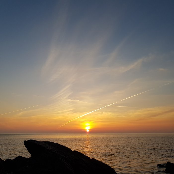 Solnedgång vid havet med strimmiga moln och stor sten i förgrunden.