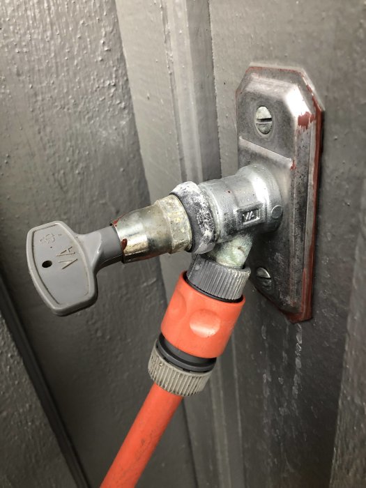 En äldre modell av vattenutkastare fäst vid en grå vägg med en orange slang kopplad till den.