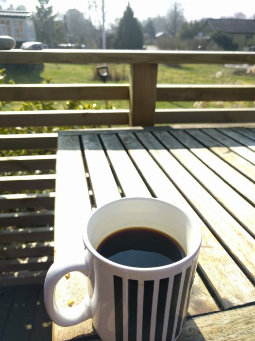 En kaffekopp på ett träbord utomhus med utsikt över en solig trädgård.