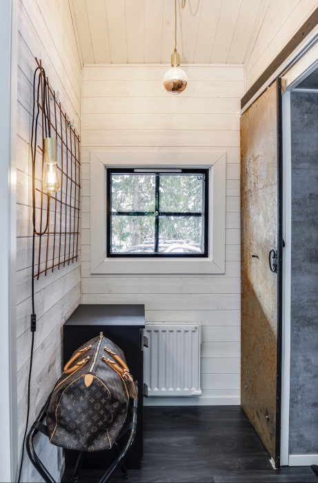 Modern hall med ljusa träväggar, designlampor och ryggsäck på stol vid öppen dörr.