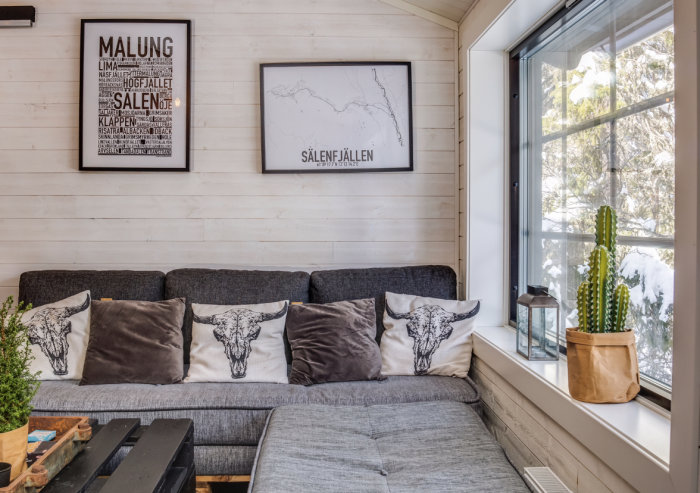 Inredningsfoto av ett mysigt hörn med grå soffa, kuddar med skalletryck, växt, kartposters på väggen och snötäckt utsikt genom fönstret.