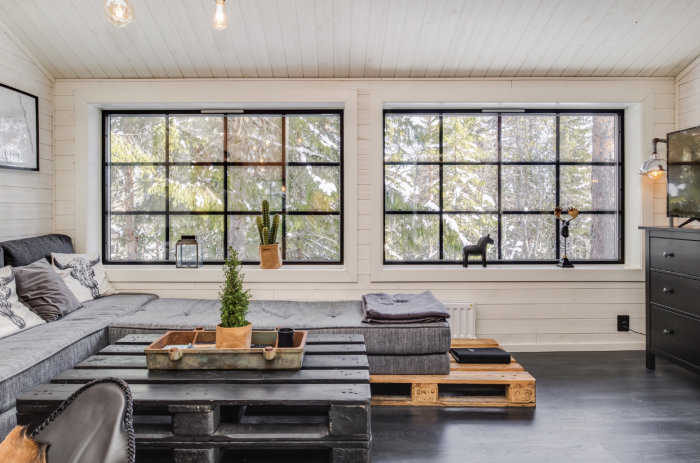 Välupplyst vardagsrum med stora fönster, skogsvy, modern soffa och rustika träbord.