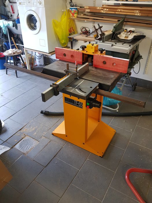 Vertikalfräs i verkstad med ytrost på bordet och tillbehör, placerad nära tvättmaskin.
