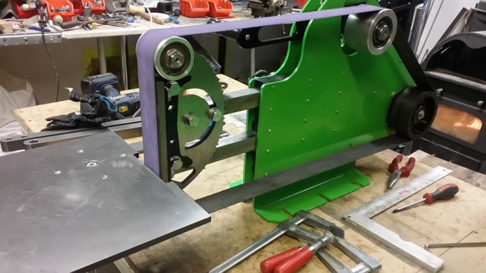 Bild på en grön och lila bandslipmaskin på ett arbetsbord med verktyg runtomkring.