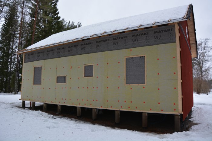 Hus under renovering med isolering och delar av röd panel på gavelsidan, på pelare i snöig miljö.