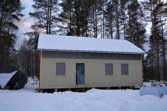 Hus under konstruktion med isolering på väggar, omgiven av snö och skog i skymningen.