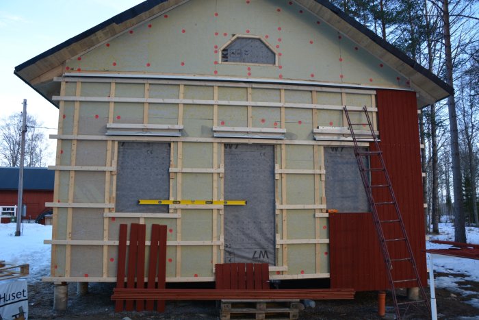 Gavel på hus under renovering med isolering, reglar och delvis monterad röd panel, stege och verktyg syns.