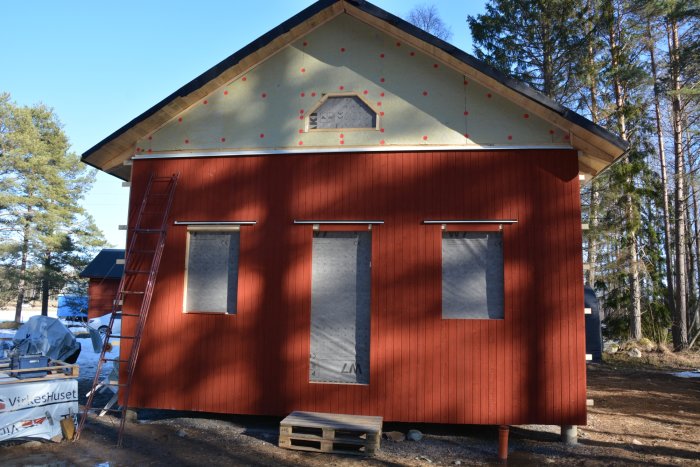 Hus med röd panel halvklart, isolering synlig, stege lutad mot väggen, skog i bakgrunden.