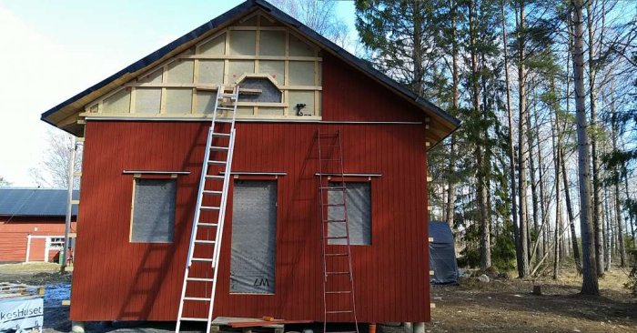 Hus under renovering med uppsatt röd panel på gaveln och stege, partiellt isolerade väggar syns.