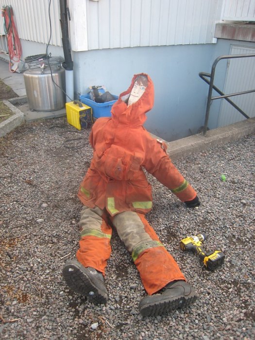 En trasig docka med arbetskläder och tejpansikte ligger på grus med en skruvdragare bredvid.