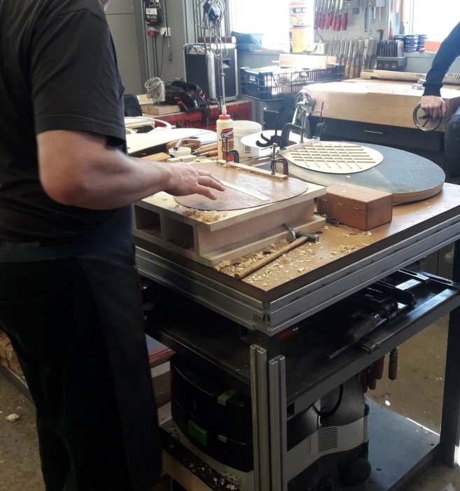 Person i verkstad hyvlar trä på arbetsbänk medan andra arbetsverktyg och projekt är synliga i bakgrunden.