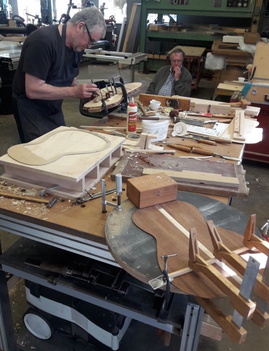 Två personer i en snickeriverkstad med olika träbearbetningsverktyg och komponenter till ett byggprojekt på arbetsbänken.