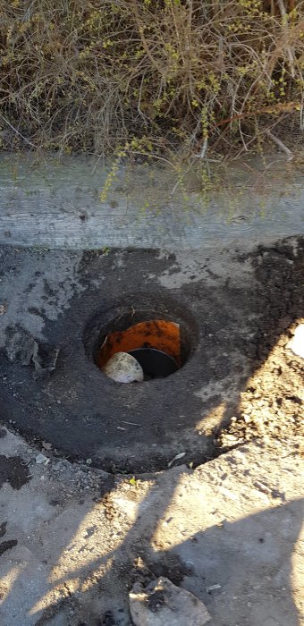 Brunn med innerdiameter 280 mm dold av stenar och löv på en uppfart nära ett hus.