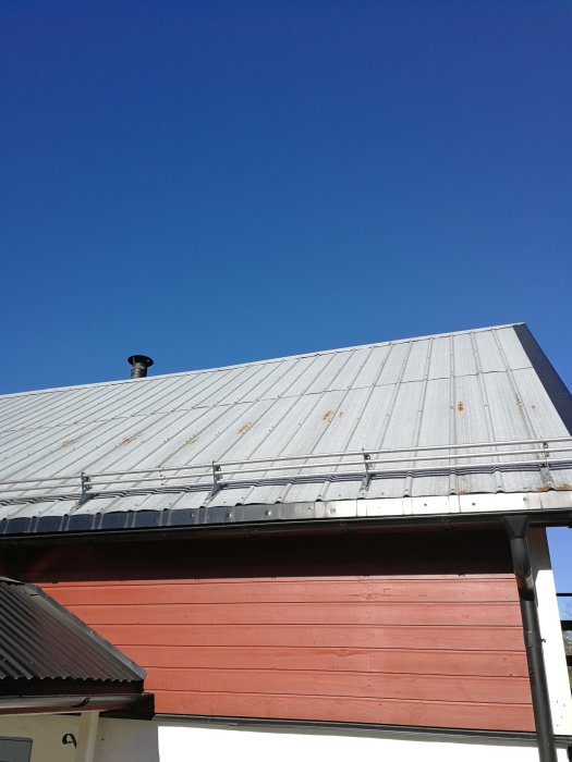 Strävt plåttak med synliga rostfläckar mot en klarblå himmel, ovanför en röd husvägg.