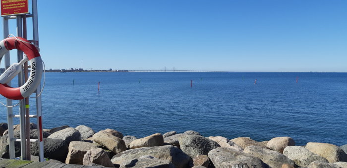 Solljus över havet med stenar i förgrunden och en bro i bakgrunden under en klarblå himmel.