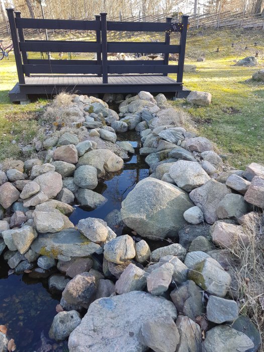 Trädgårdsdamm med stenbädd och galler under småsten, med träbro över vattenytan.