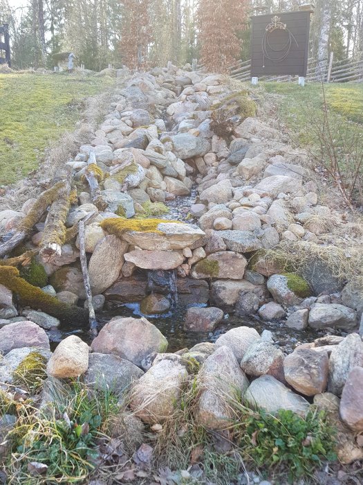 En stenbelagd bäck med ett osynligt säkerhetsgaller och omgivande grönska i en trädgård, ej i full blom.